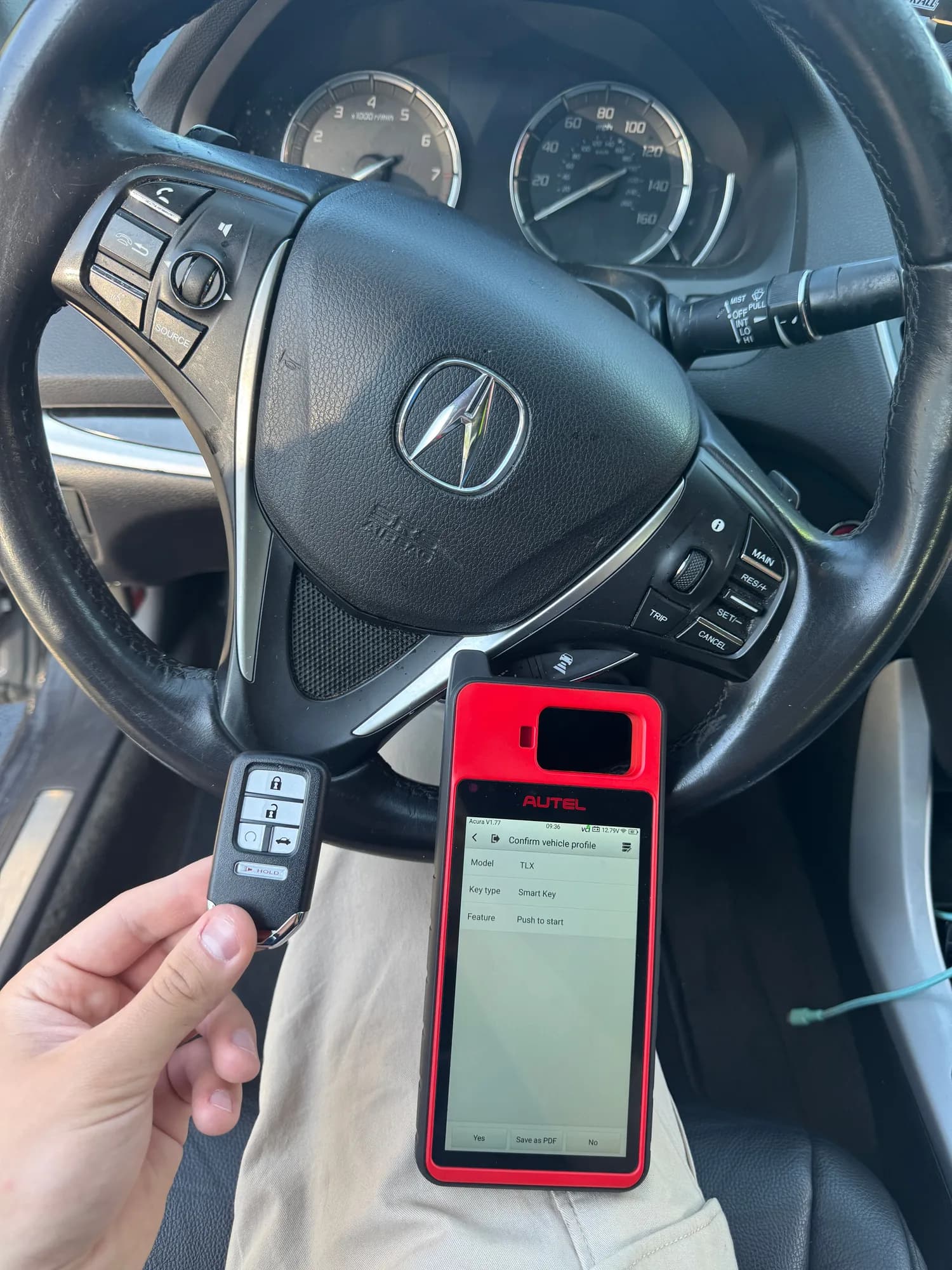 A hand holding an Acura smart key in front of the steering wheel of a TLX while a programming tool on the technician's knee shows the vehicle profile it has identified