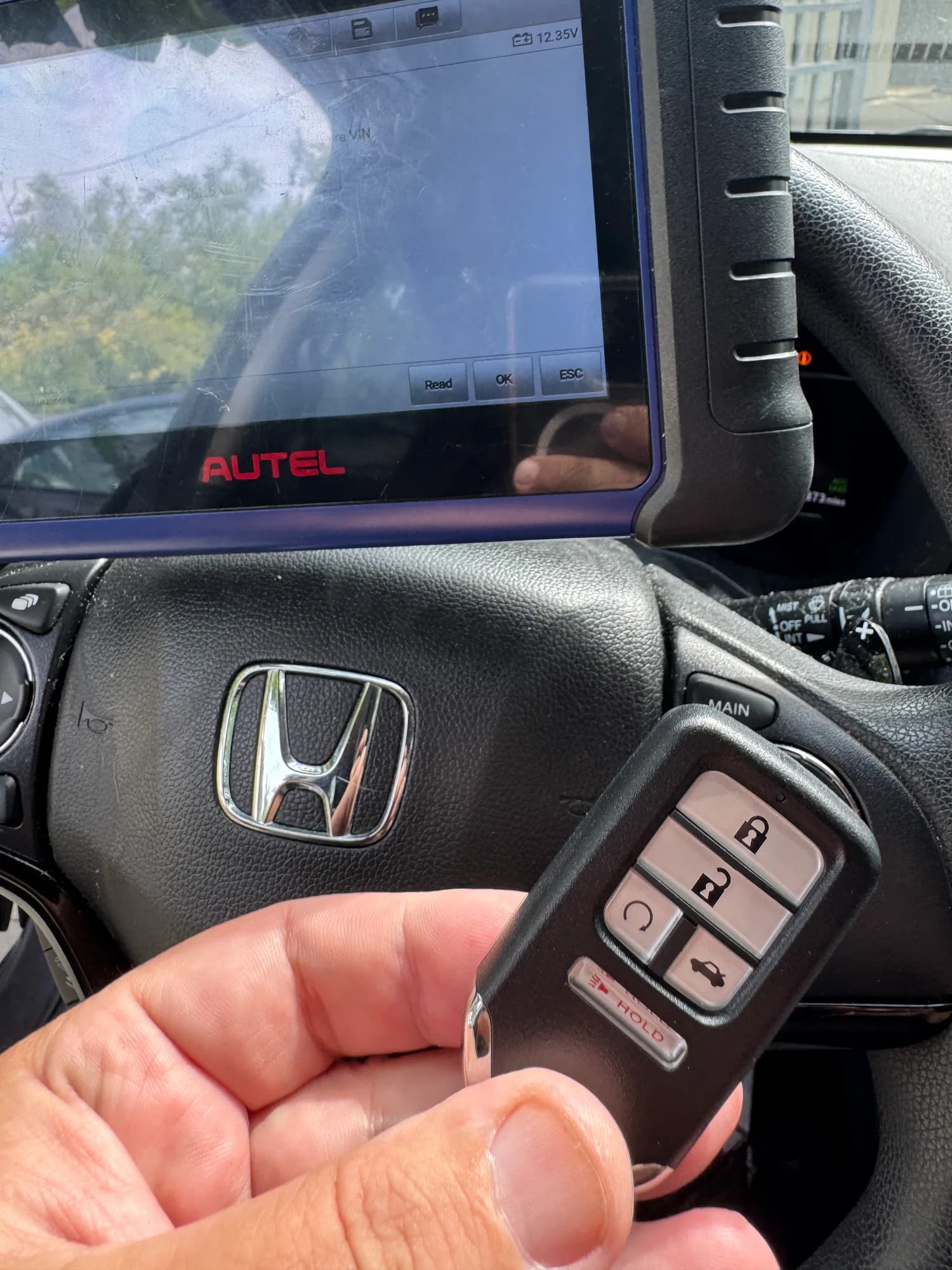 A Honda smart key held in front of the steering wheel while a programming tablet plugged into the car shows the key-learning screen