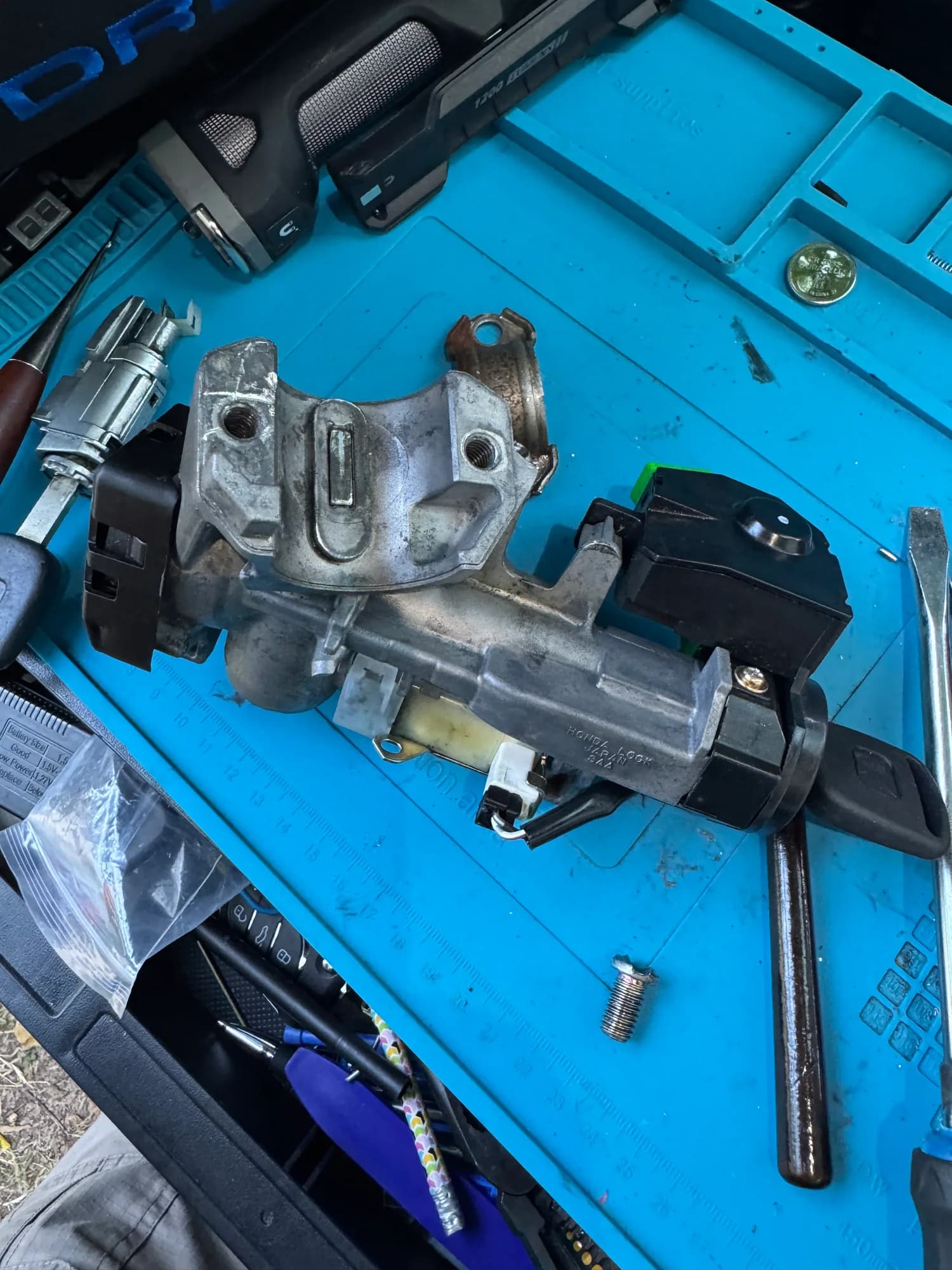 An ignition lock cylinder and steering column lock assembly taken apart on a work mat, surrounded by picks, screwdrivers, a battery tester and cut transponder keys