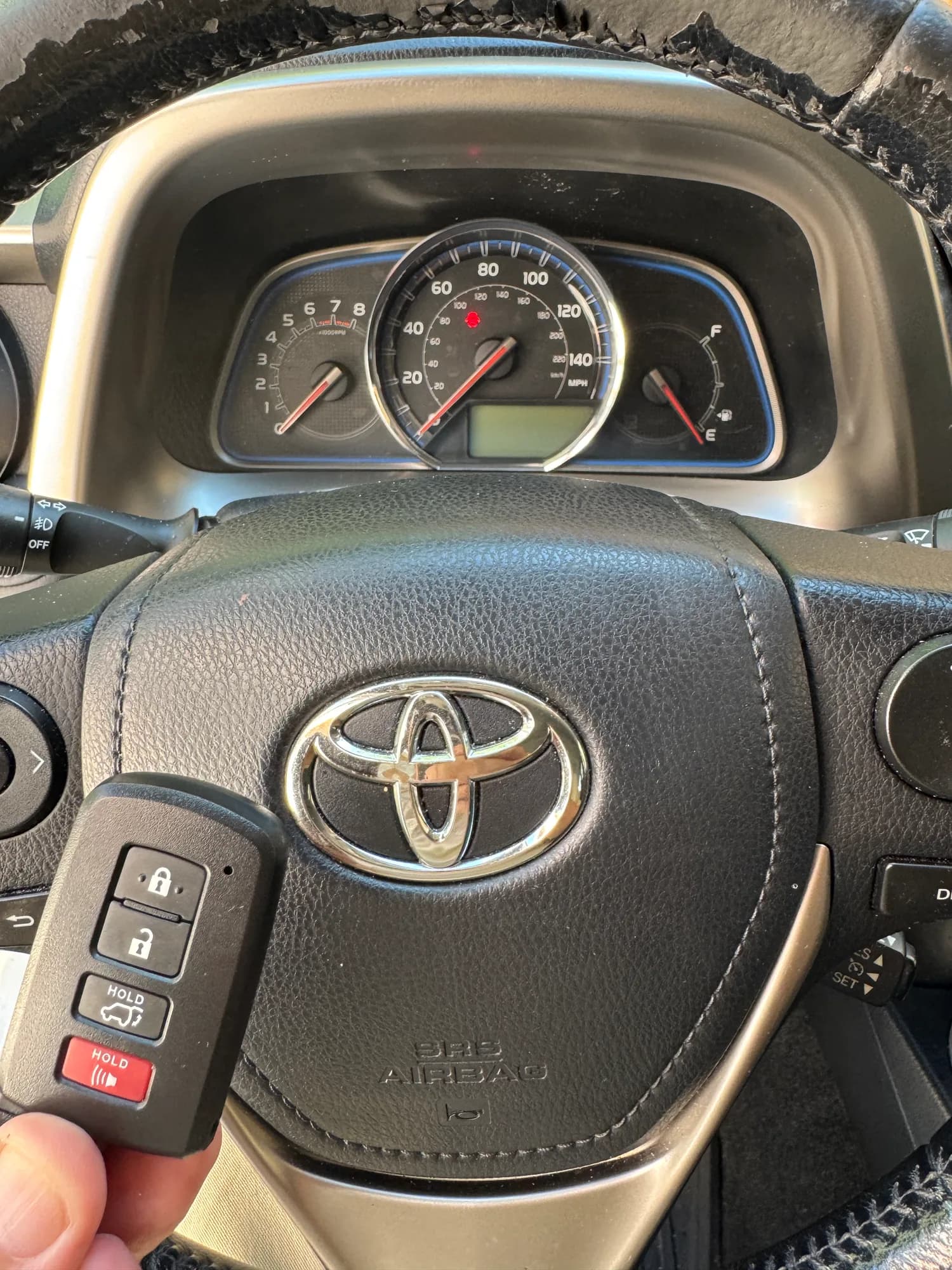 A four-button Toyota smart key held in front of a Toyota steering wheel and instrument cluster