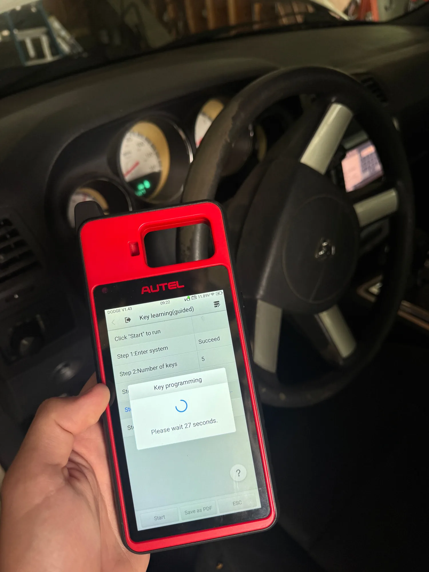 An Autel key programmer held in front of a Dodge steering wheel, its screen on the Dodge key-learning procedure with step one marked Succeed and a 27-second wait running