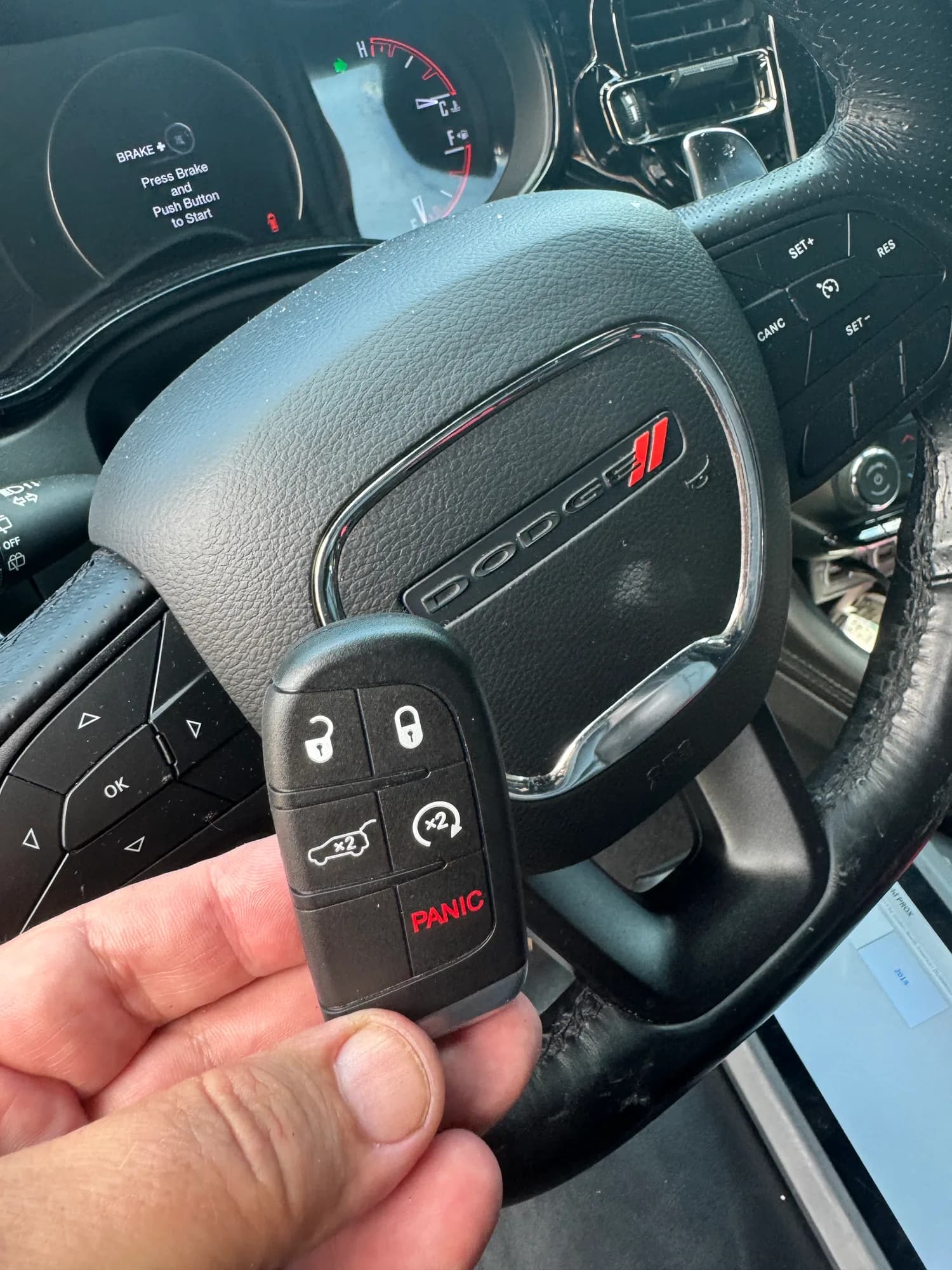 A five-button Dodge smart key held in front of the steering wheel of the car it was made for, with the dash reading Press Brake and Push Button to Start
