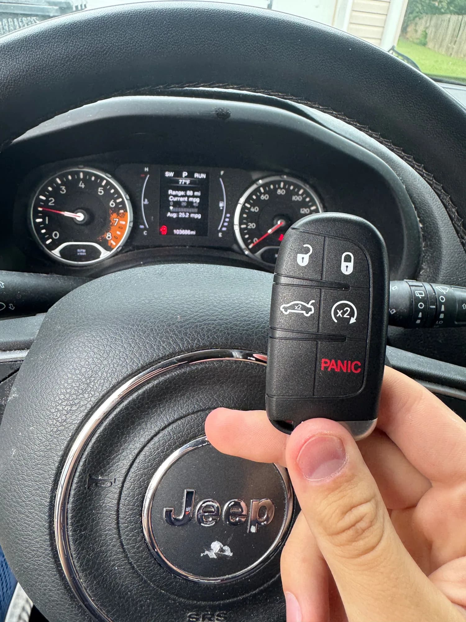 A five-button Jeep smart key held in front of a Jeep steering wheel, the dash showing the vehicle in park and running