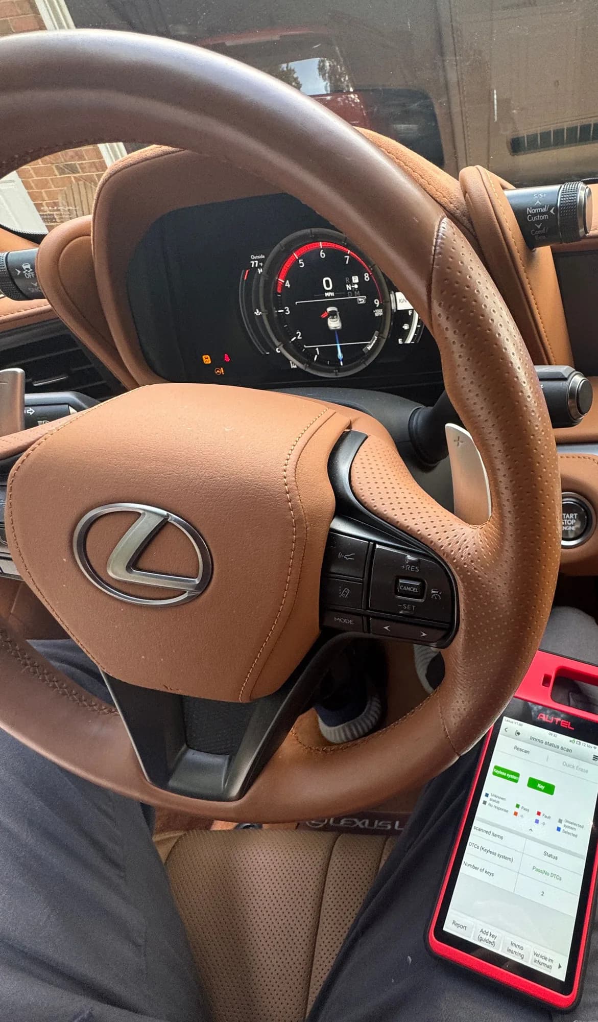 A programming tool resting against the tan leather steering wheel of a Lexus, its screen reading Immo status scan with the keyless system passing and the number of keys registered to the car showing two