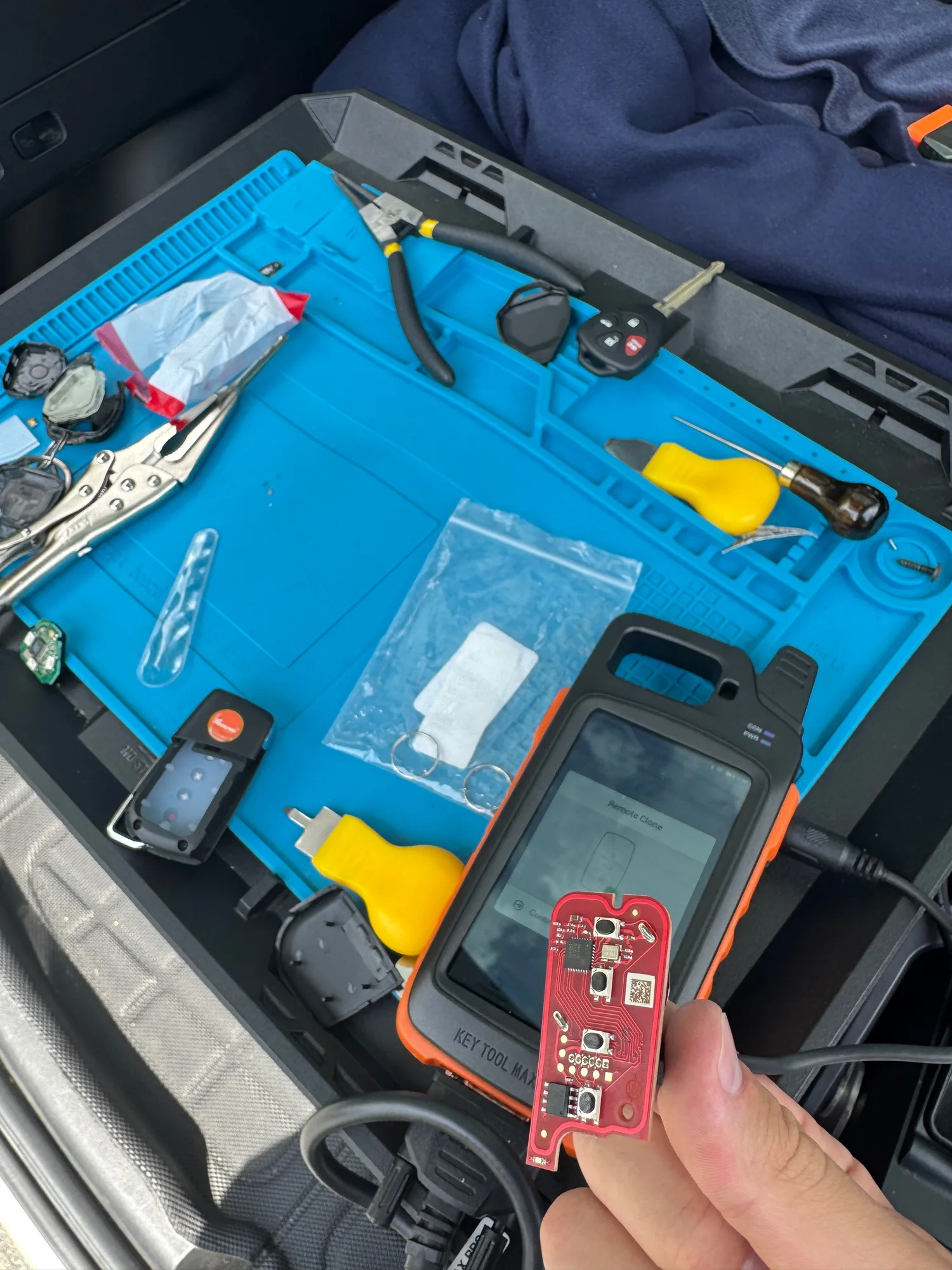 A work mat laid out in the back of a service vehicle with locking pliers, screwdrivers, key shells and a remote circuit board held in front of a key programming tool